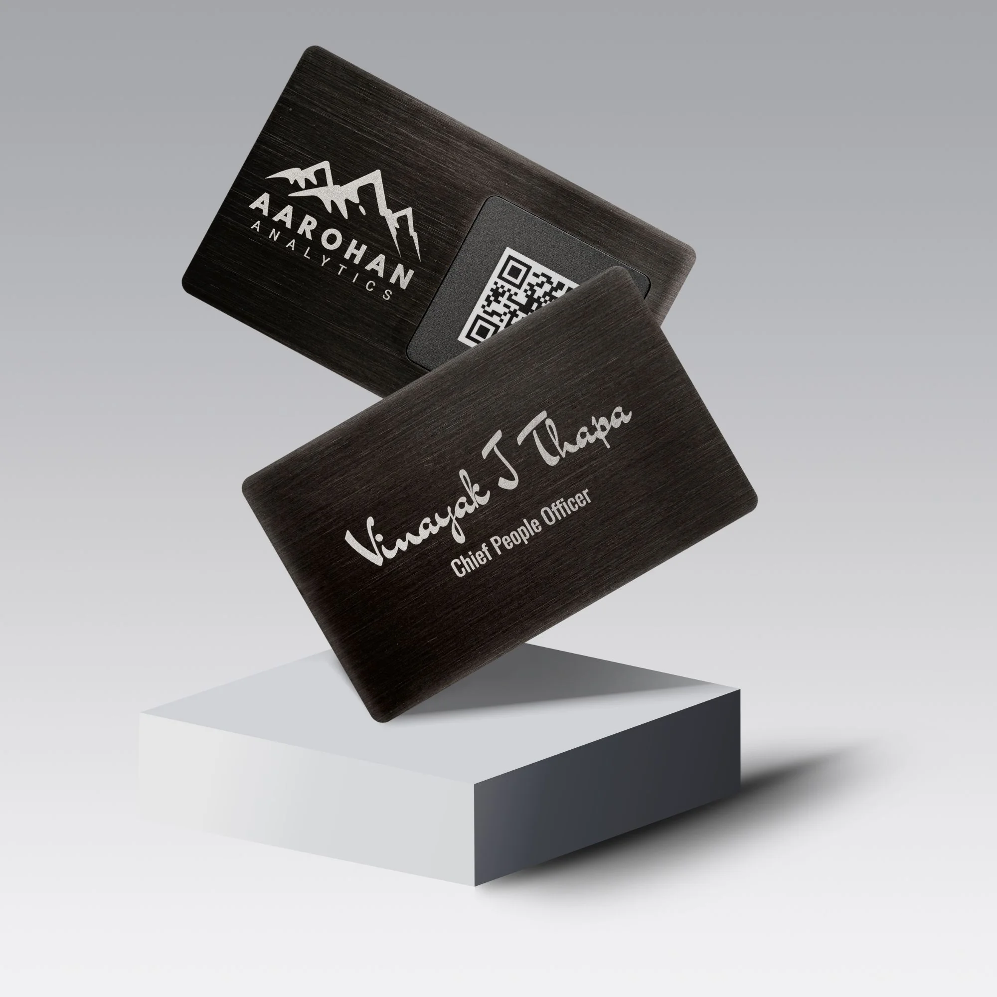 Two sleek gun black metal cards with engraved text and a QR code, displayed on a white pedestal.