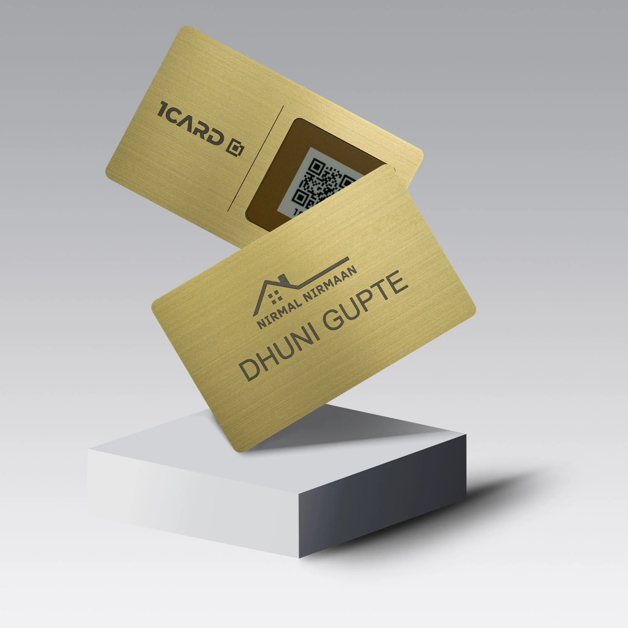 Two gold metal business cards with engraved text and a QR code, displayed on a white pedestal.