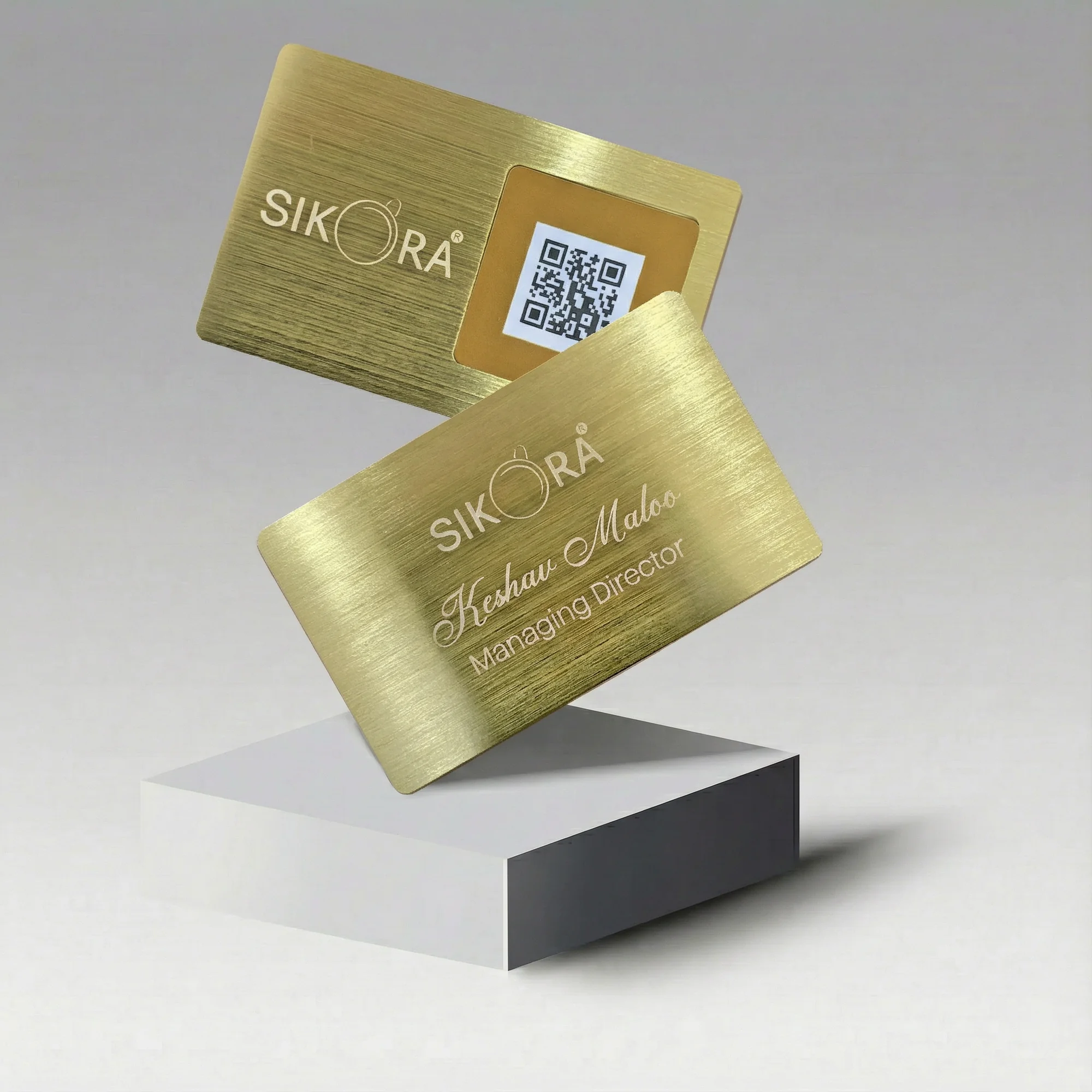 Two gold metal business cards with engraved text and a QR code, displayed on a white pedestal.