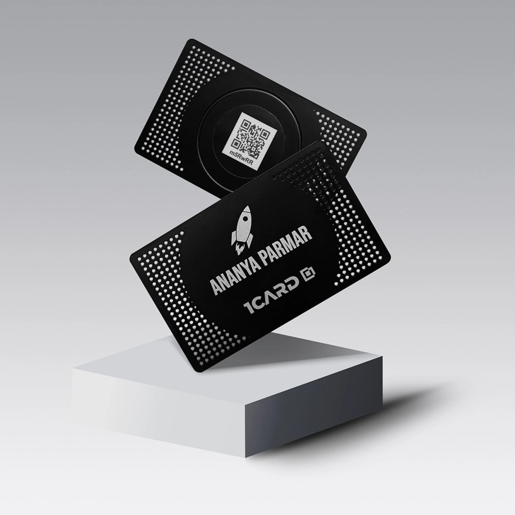 Two black metal cards with perforated edges, one displaying a QR code and the other showing "ANANYA PARMAR" and "1CARD" logos, on a gray gradient background.